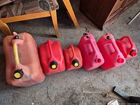 Six assorted plastic gas cans lined on floor showing condition, colors, and presence or absence of caps