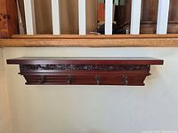 Front view of wall-mounted wooden shelf showing carved floral design and five metal hooks
