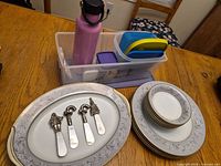 Overview of Noritake china, cheese spreaders, water bottle, plastic containers on table