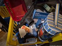 Overview of yellow crate containing iron pry bar, Shift boxed item, vacuum nozzle, striped tin canister, glue bottle, dustpan and basket