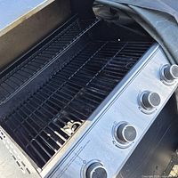 Open grill showing interior grates, control panel with four knobs, partial view of cover