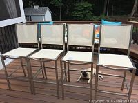 Four stainless steel frame outdoor bar height chairs with ivory mesh seats and backs, positioned side by side on a wooden deck with a pug lying nearby.