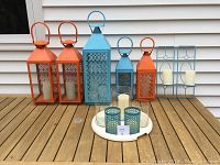 Photo of five orange and turquoise metal lanterns arranged on a wooden tabletop, with a white metal tray holding candle holders in front. Some lanterns have candles inside and some have missing glass panes.