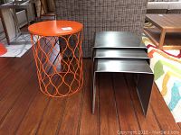 Orange lattice-style metal side table next to three stacked stainless steel nesting tables, viewed from side on wooden floor.