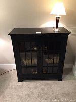 Front view of black painted wood cabinet with glass-paneled doors and lamp on top.