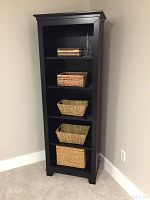 Front view of tall black painted wood bookcase with five shelves, each holding woven baskets