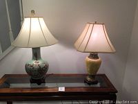 Pair of two decorative table lamps on a wooden table: taller blue floral ceramic lamp with finial on left, shorter beige ceramic lamp on right
