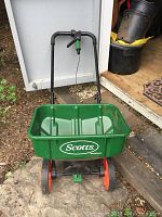 Green Scotts fertilizer spreader with black handle and red wheels, standing on yard gravel with door and black container in background.
