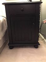 Front view of the black painted wood night stand showing drawer and paneled door with knobs, set on bun feet.
