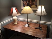 Photo of three table lamps arranged on a wood table against gray walls showing their relative heights and shade designs.