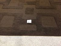 Close-up of chocolate brown rug showing texture and geometric rectangular pattern with some wear.