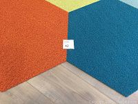 Close-up of two hexagonal carpet tiles in orange and turquoise colors placed side by side on a wood floor.