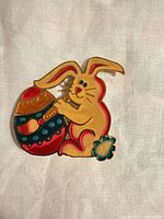Front view of enamelled metal bunny holding decorated egg brooch