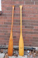 Both wooden canoe paddles leaning against brick wall showing full length and color difference