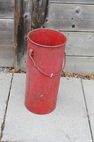 Full view of red milk can standing upright on outdoor pavers, showing handle and wear
