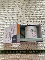 Mug in open gift box showing text “merry” and green handle, measuring tape for scale