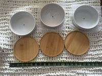 Three white ceramic planters with drainage holes and three bamboo saucers, tape measure for scale