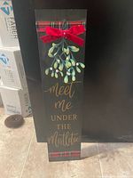 Front view of vertical black wooden sign showing metal mistletoe, red bow, plaid trim, gold text