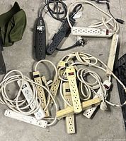 Top-down view of all twelve surge protectors piled on concrete floor, showing mix of colors and outlet counts