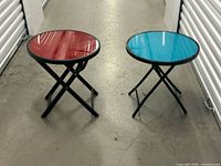 Both round glass folding tables side by side, showing red and turquoise tops and black frames
