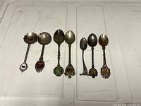 Front view of seven loose souvenir spoons showing enamel crests and varying bowl shapes