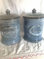 Front view of both blue-grey ceramic flour canisters with lids and raised “Flour” plaques