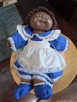 Front view of Cabbage Patch Kids doll in blue dress and white apron lying on table