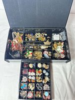 Open jewelry organizer showing upper and lower trays filled with assorted brooches, earrings, necklaces, and rings