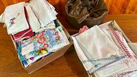 Overview of two boxes containing assorted folded linens and hankies plus brown cylindrical container