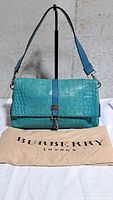 Front view of turquoise embossed croc Burberry flap bag on stand with dust bag