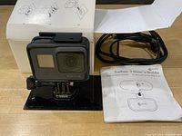 Front and top view of GoPro camera on mount with cable, manual, box visible