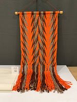 Front view of orange brown beige woven wall hanging on wooden rod with fringe, letter partially visible