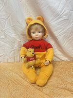 Front view of porcelain doll wearing Pooh suit holding plush bear