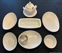All seven white serving pieces laid out including soup tureen with lid on underplate, various bowls and rectangular dish