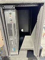 Front view showing rack-mounted QSC amplifier and empty bay inside carpeted cabinet