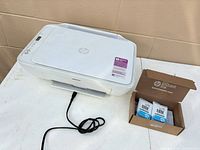 HP white all-in-one printer with power cord and boxed cartridges on table