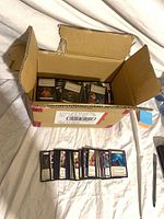 Open cardboard box containing stacks of Flesh and Blood trading cards with additional cards lined up in front