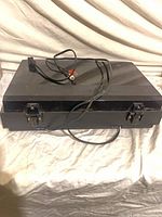 Turntable with lid closed, power cord and RCA cable on top, front latches visible