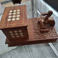 angled view of carved wood cigarette dispenser showing bird lever, floral base, inlaid dots