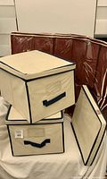 Three beige fabric cubes with navy trim stacked, maroon under-bed organizer behind