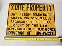 Front view of yellow and black traffic sign with full text visible and tape measure indicating size