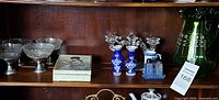 Shelf showing metal bowls, porcelain candleholders with glass tops, sewing box, part of green pitcher