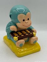 Front view of turquoise ceramic monkey piggy bank holding abacus on yellow base