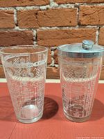 Both glass cocktail shakers, one with metal lid attached, showing printed recipes and ounce scale