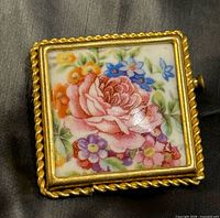 Front view of square ceramic brooch with floral painting and gold-tone rope border