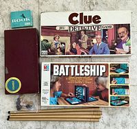 Lot overview showing game boxes for Clue, Battleship, Rook, Scrabble box lid, crokinole sticks and discs