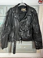 Front view of FMC black leather motorcycle jacket on hanger showing asymmetrical zipper, belt, lapels, pockets