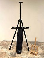 Full view of one metal tripod easel, carry bag leaning against it, wooden tabletop easel beside