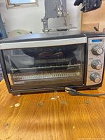 Front view of Black & Decker toaster oven showing glass door, control panel with four knobs, interior rack and tray, attached power cord