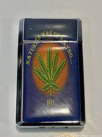 Lighter closed showing blue enamel, cannabis leaf graphic, chrome frame
